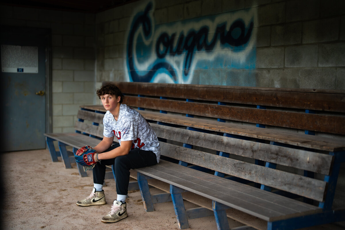 High school senior boy sitting in a dugout with a baseball glove.