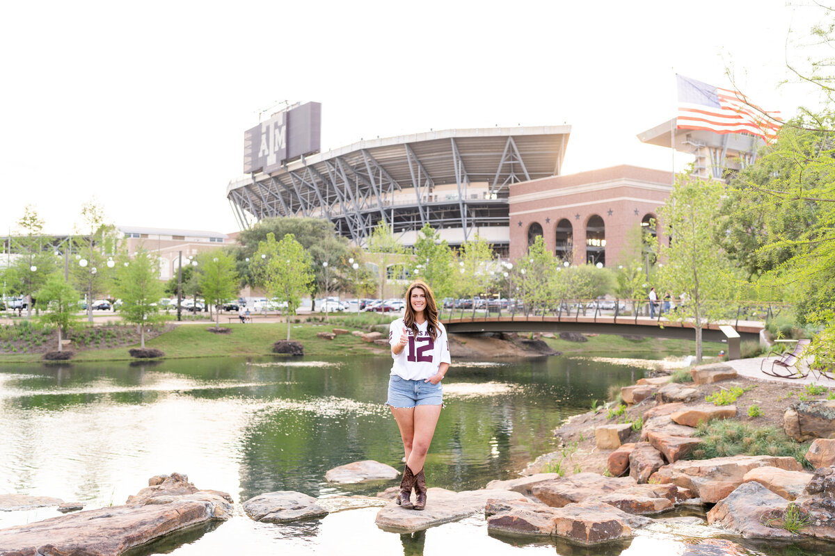 Texas A&M senior girl standing on rocks with hand in pocket and thumbs up wearing white jersey in Aggie Park in front of Kyle Field