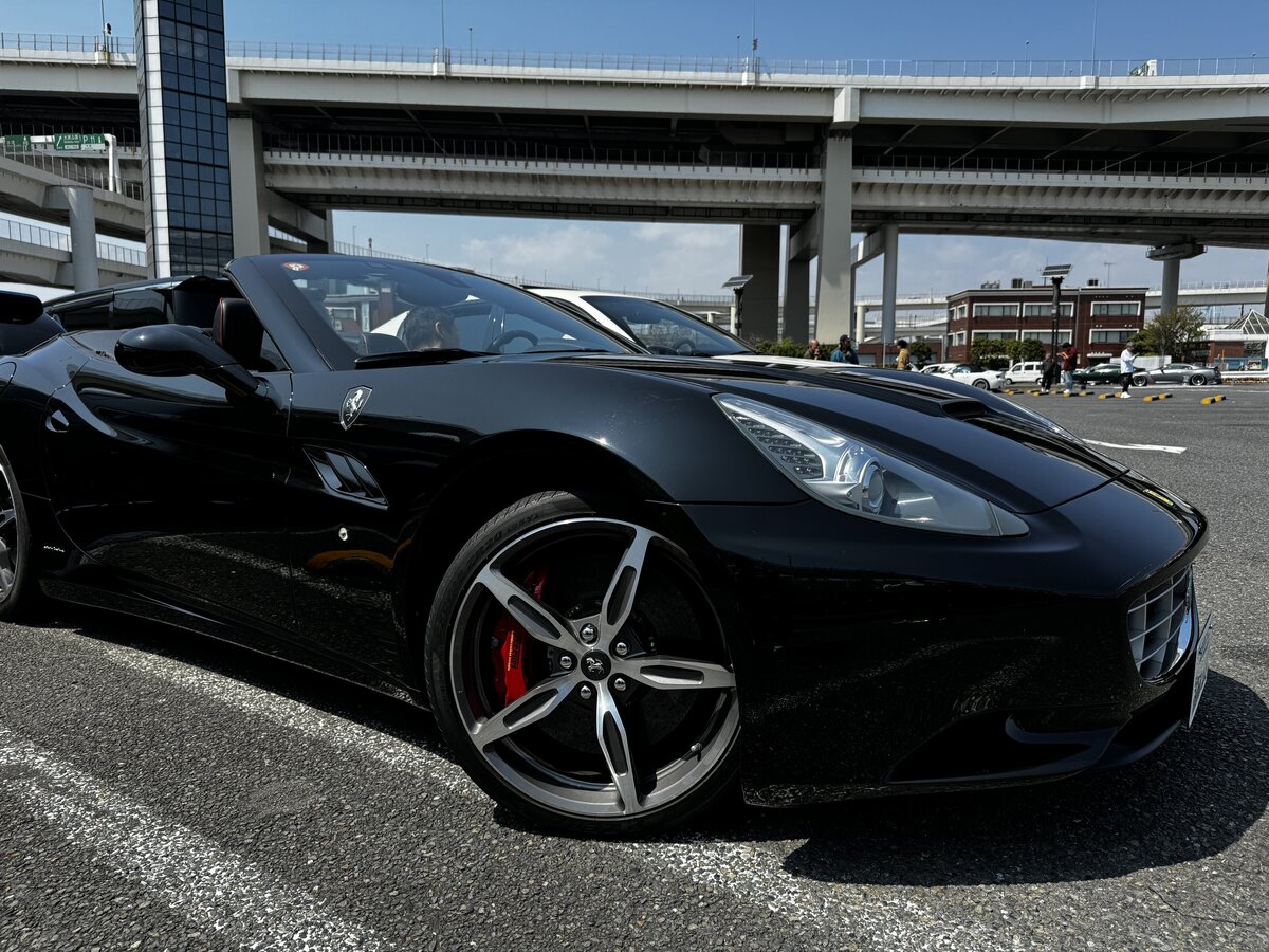 A sleek black Ferrari California captured from a low front angle, parked at Daikoku Parking Area with highway overpasses in the background.