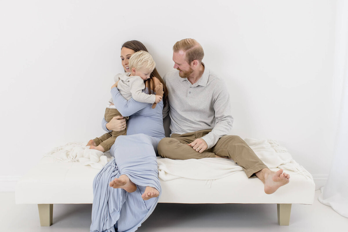 A family of three sits together on a white sofa against a plain background. The mother in a long blue dress holds a young child who is hugging her. The father, in a gray shirt, sits beside them, smiling at the child.