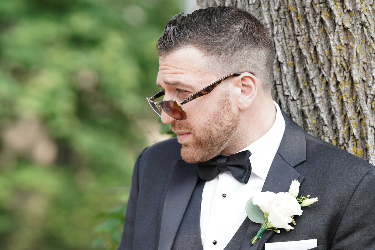 A close-up of the groom leaning casually against a tree while wearing sunglasses. This image captures a relaxed and stylish moment, highlighting the groom’s modern, confident look and adding a touch of personality to his wedding portraits.