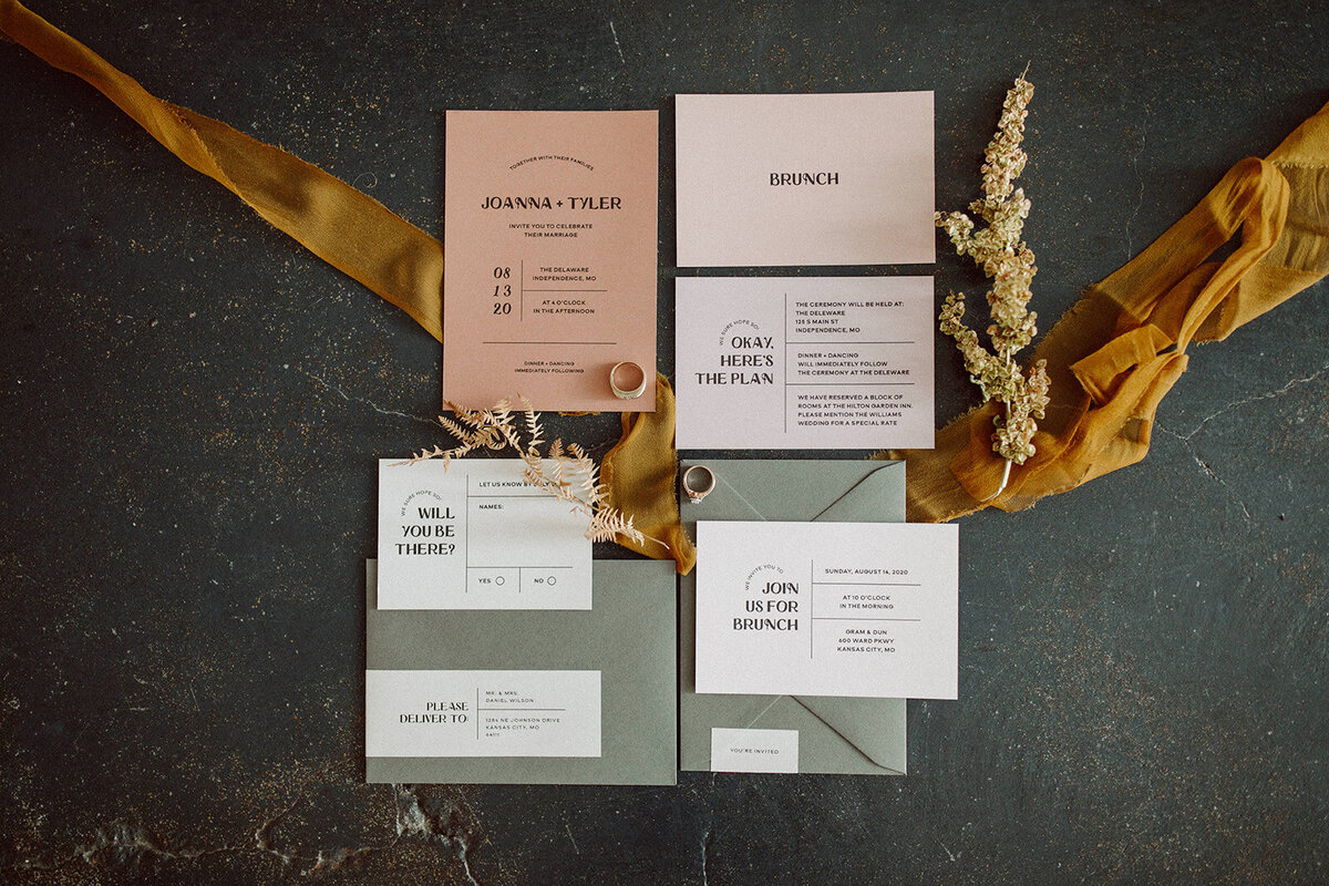 Off-white, peach and mauve-colored wedding stationery with black font placed next to two wedding bands atop ribbon.
