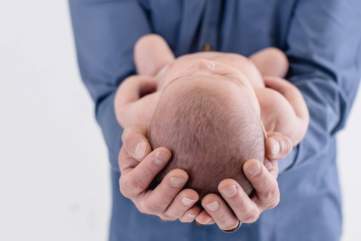 A person in a blue shirt gently cradles a newborn baby, focusing on the babys head and the persons hands. The background is softly blurred.