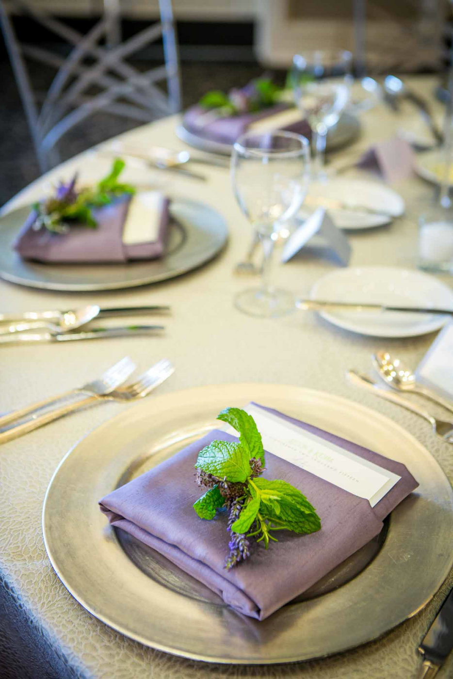 place setting with silver charger plate, purple napkin, and bunch of herbs as napkin treatment