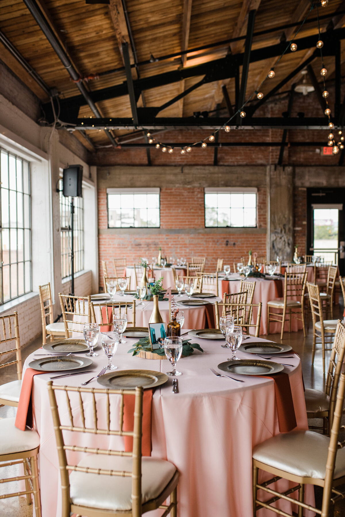Room shot of a wedding reception at the Hickory Street Annex in Dallas, Texas. Multiple round tables can be seen with pale pink tablecloths, dark brown plates, cutlery, glasses, and a centerpiece containing a vase with flowers, a  vase with a light, a framed photo, and a table number.