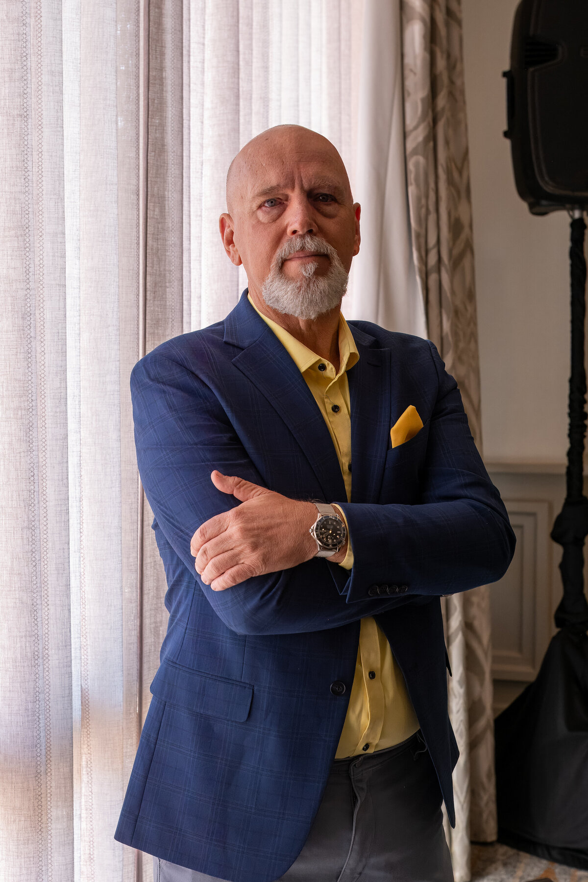 Business portrait of a man posing with crossed arms near a curtain, wearing a blue blazer, photographed by Vyrl Photo in Tucson.