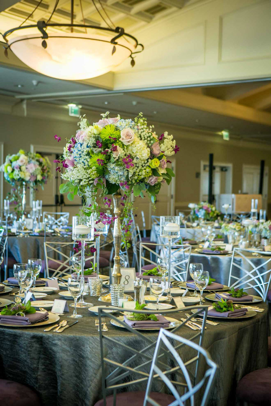 large centerpiece of blue hydrangea, purple roses, green chrysanthemums, , and trailing purple clematis, with floating glass candle holders on silver linen and silver Chameleon chairs