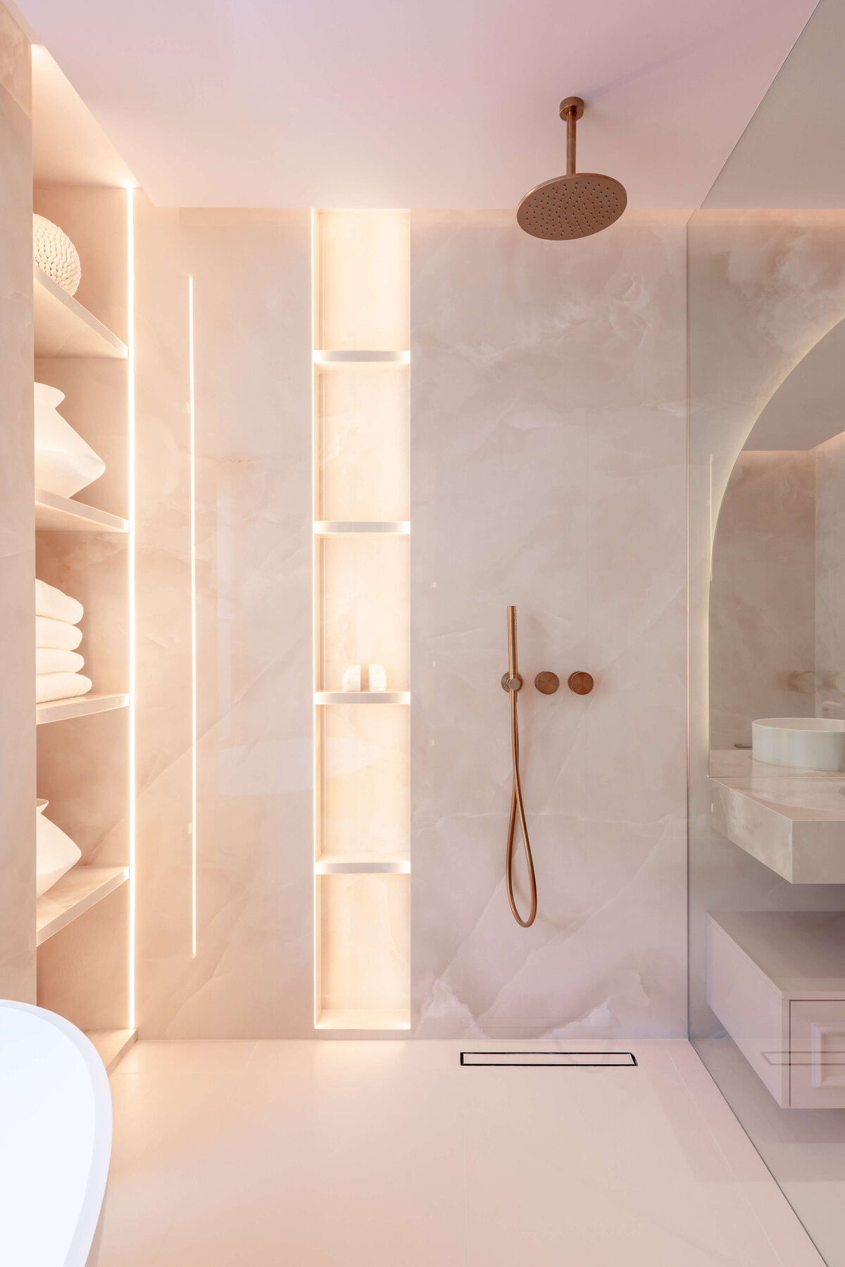 Large open shower with light pink marble walls and rose gold hardware. There is built in shelving that is backlit to emit a soft glow.