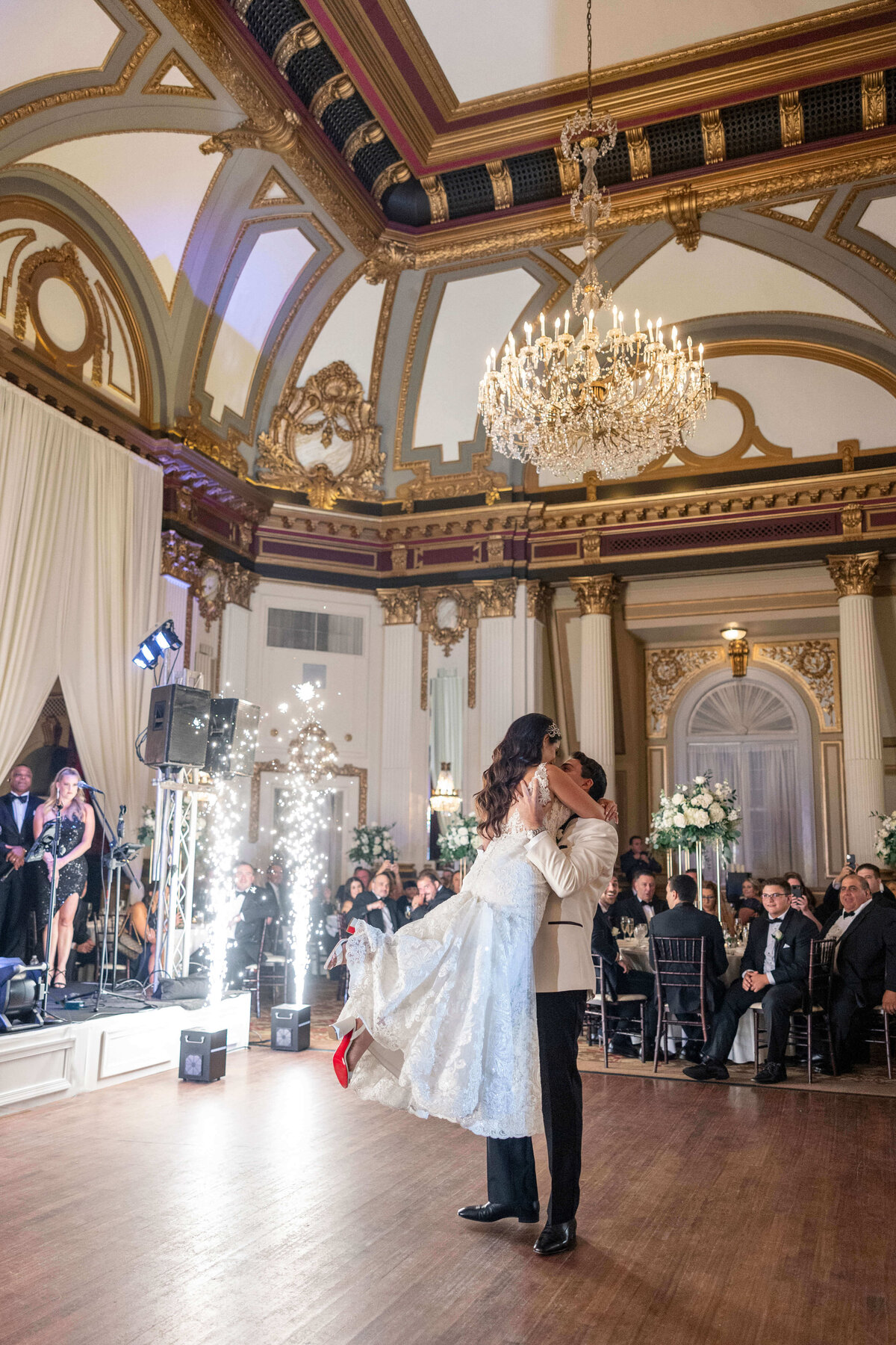 A couple enjoys their first dance in an opulent ballroom with a grand chandelier. The groom lifts the bride, who is wearing a white gown. Guests watch from elegantly decorated tables, and a backdrop of fireworks adds to the celebratory atmosphere.