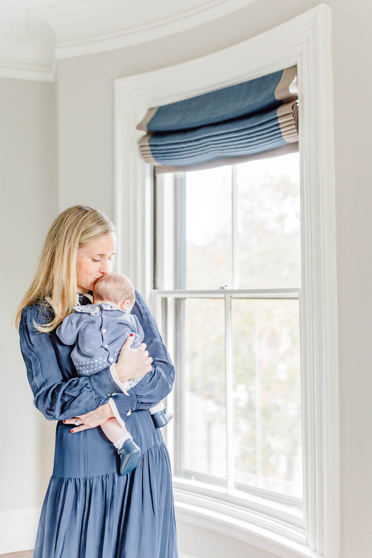 Mom in a blue dress holding her sleeping baby next to a window in Boston