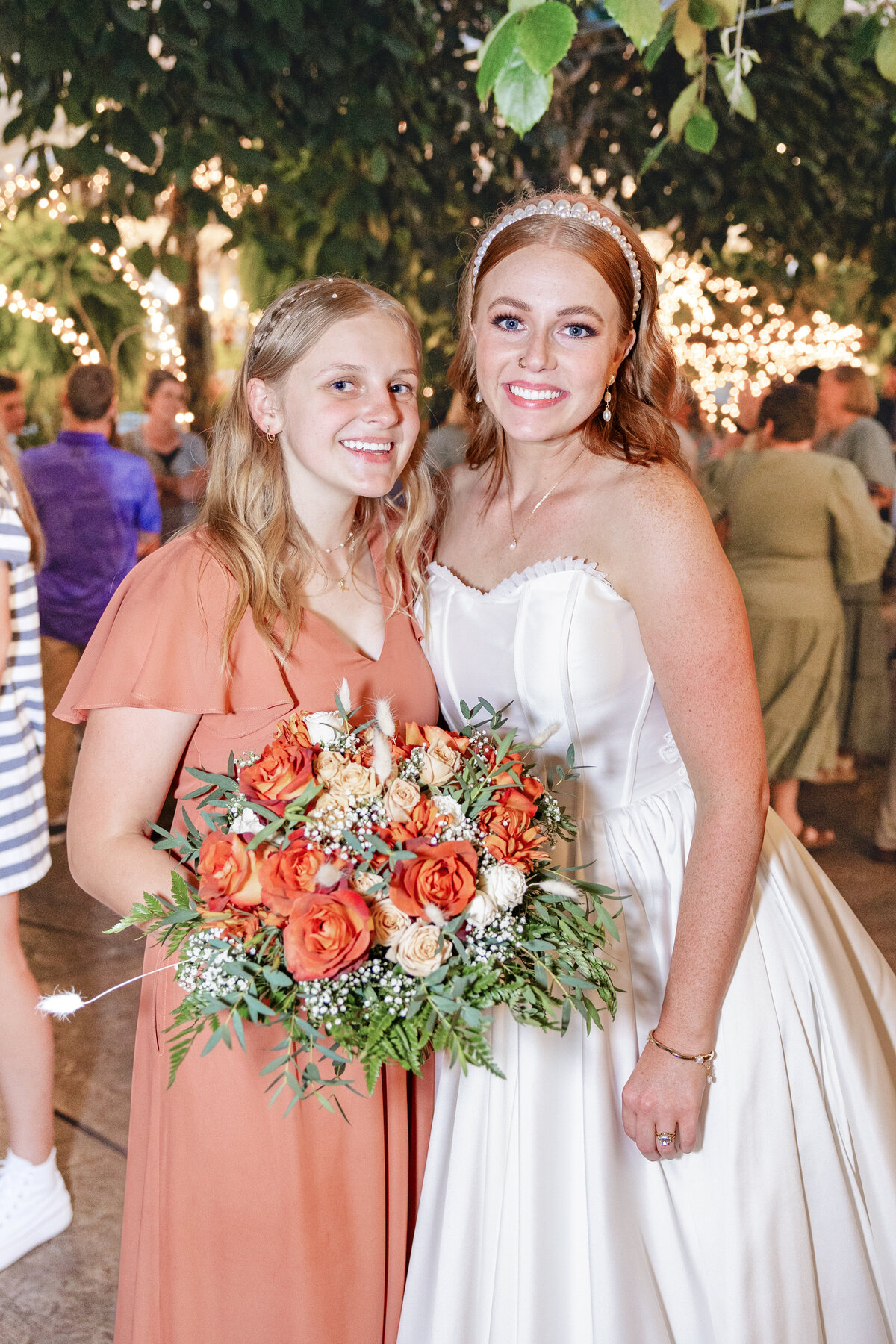 A bride in a white wedding gown and a bridesmaid in a burnt orange dress smile together, holding a vibrant bouquet of orange and cream roses, surrounded by warm lights and wedding guests at Le Jardin in Sandy, Utah.