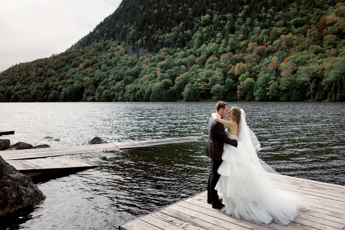 The bride and the groom are touching noses while standing on a dock facing the mountains and sea at The Ausable Club, New York.
