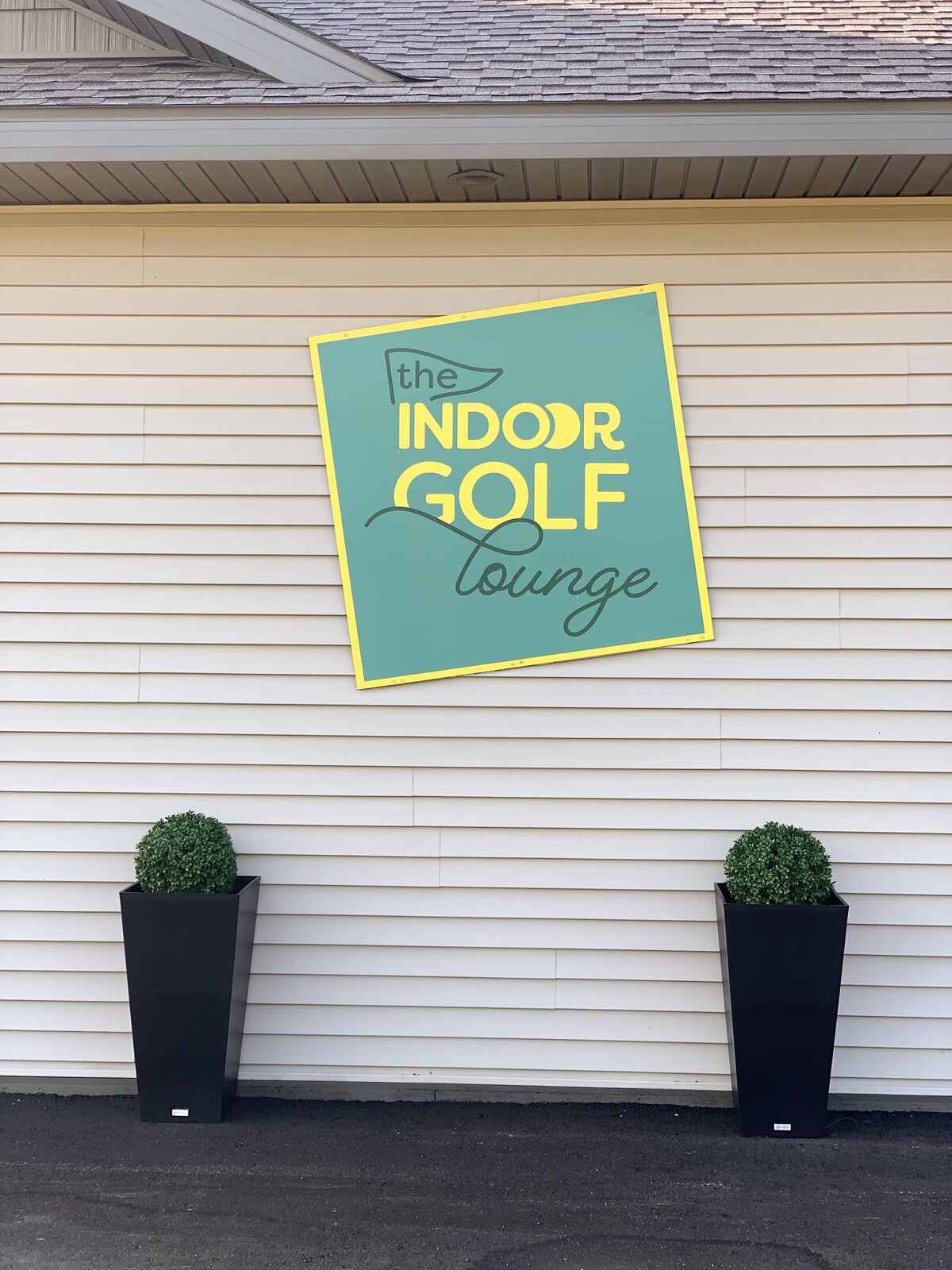 A large sign printed with The Indoor Golf Lounge logo is mounted on an exterior wall between two planters