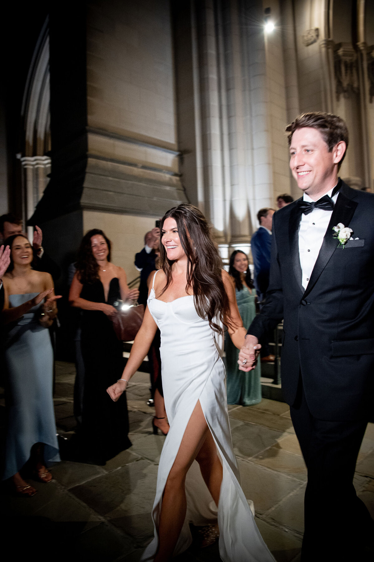 This enchanting moment was captured at the stunning Washington National Cathedral in Washington D.C. by the talented photographer Twah Dougherty and videographers, Current Films. A happy couple walks hand in hand at their wedding, with the bride looking radiant in a white dress with a high slit, accentuated by the groom's dapper look in a black tuxedo paired with a bow tie. Surrounded by guests in elegant attire, they are embraced by applause within the grand and beautifully illuminated venue.