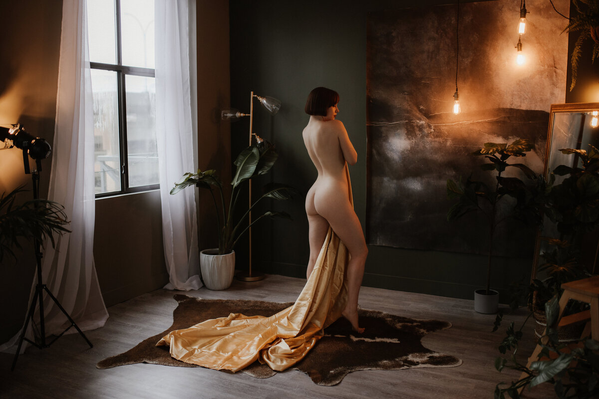 A naked woman holding a gold bedsheet poses in front of a window in the Victoria, BC boudoir studio.