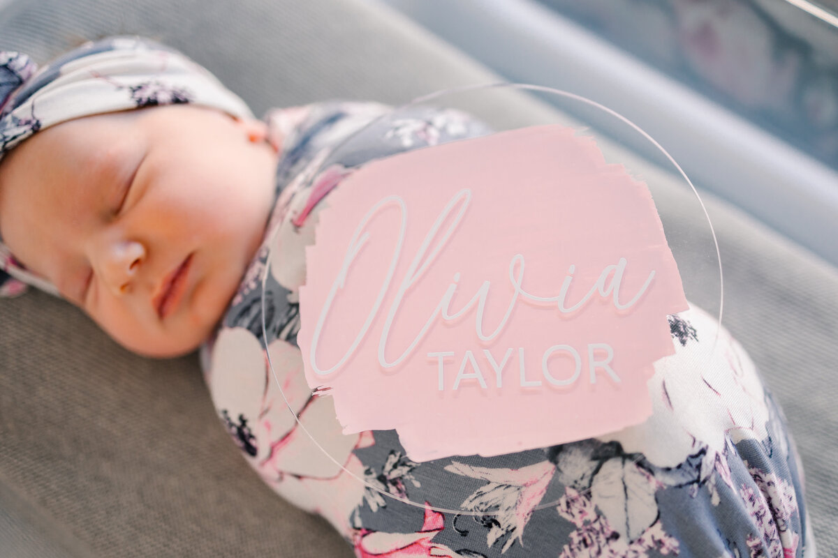 Newborn baby girl asleep on a dark gray blanket. She is wrapped in a cozy blanket and adorned with a blue headband featuring white flowers.