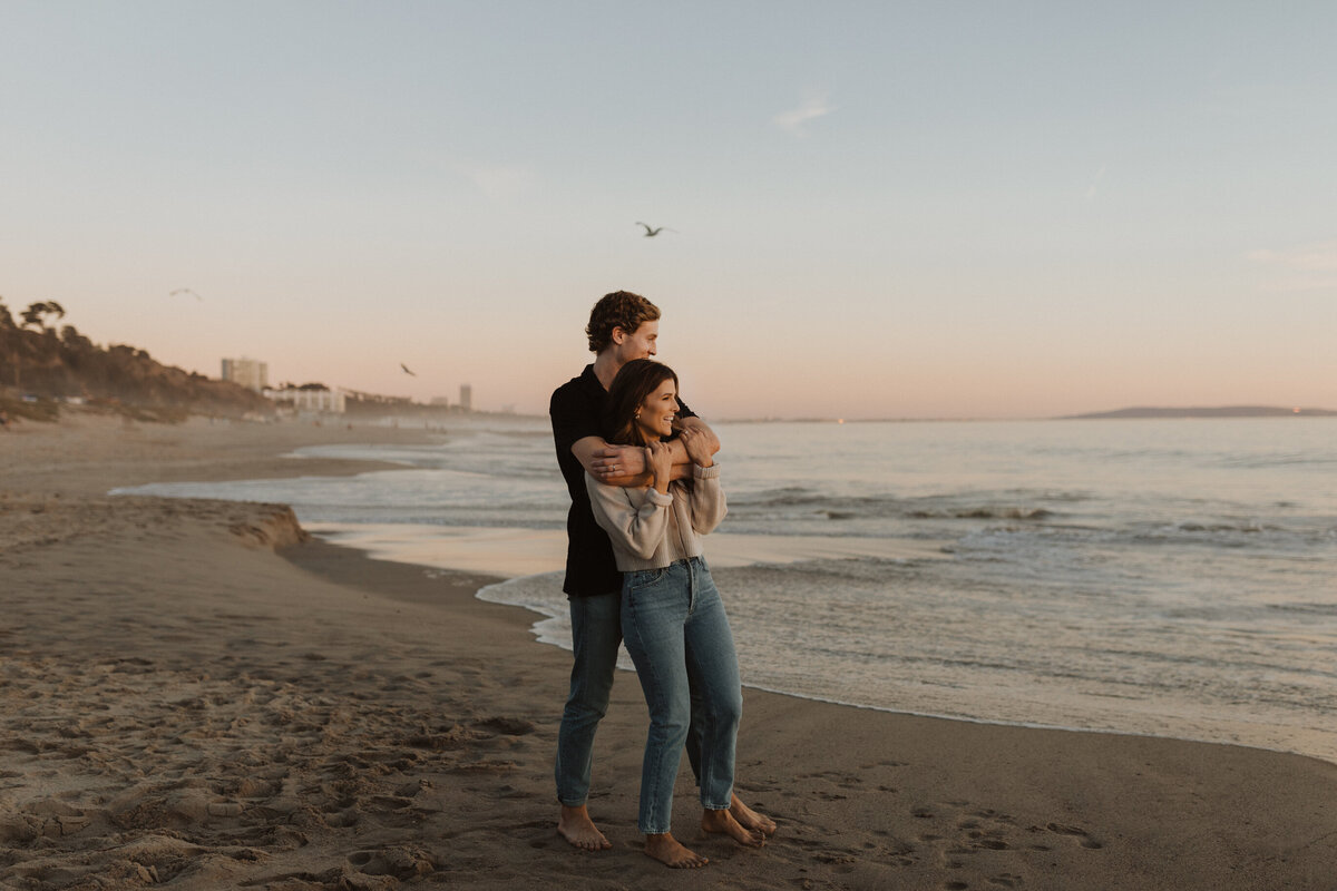 Man hugging woman from behind while they stand on the beach in front of sunset