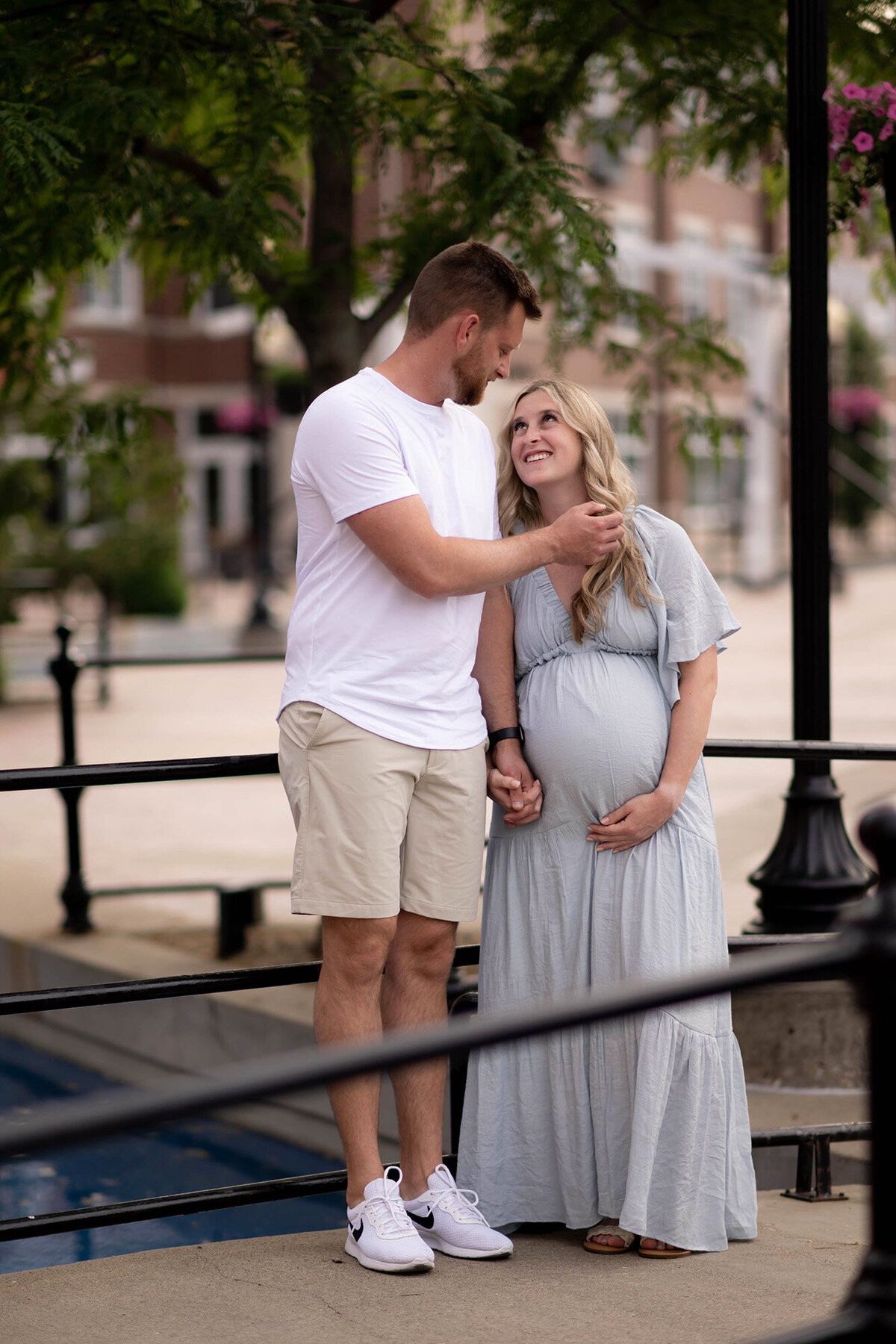 A happy pregnant couple stands on a bridge over water holding hands smiling at each other