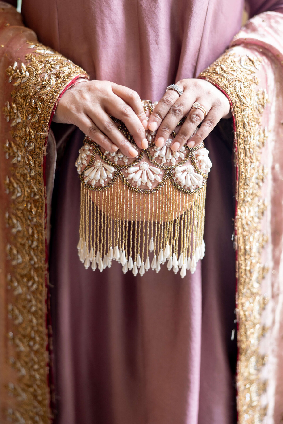 A person wearing an embroidered pinkish-brown garment holds an ornate clutch decorated with pearls and gold beaded tassels. The garment features intricate gold embroidery on the sleeves.