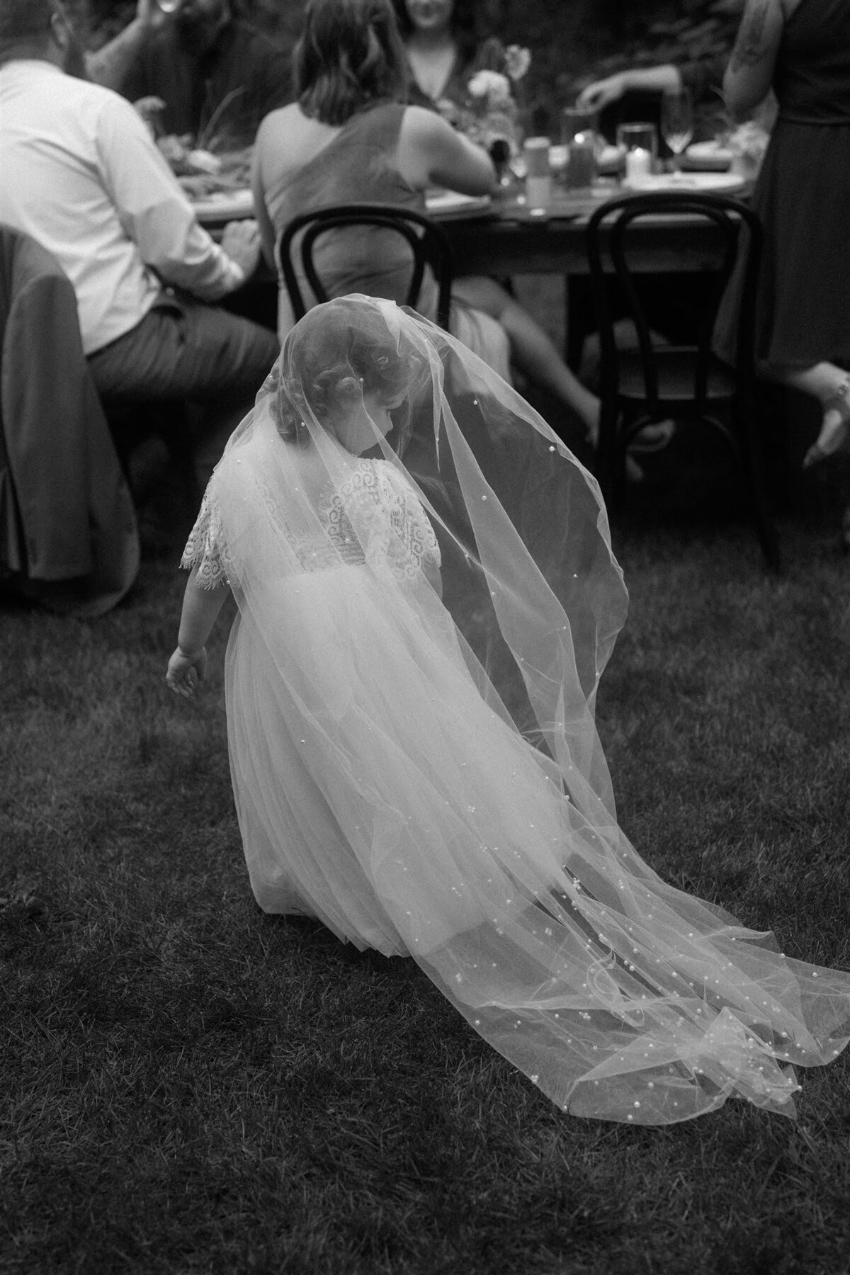 Black & white photo of flower girl walking through intimate wedding reception in the bride's veil!