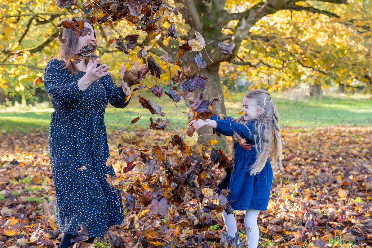 A woman and a young girl joyfully throwing autumn leaves in a park with trees displaying fall colors.