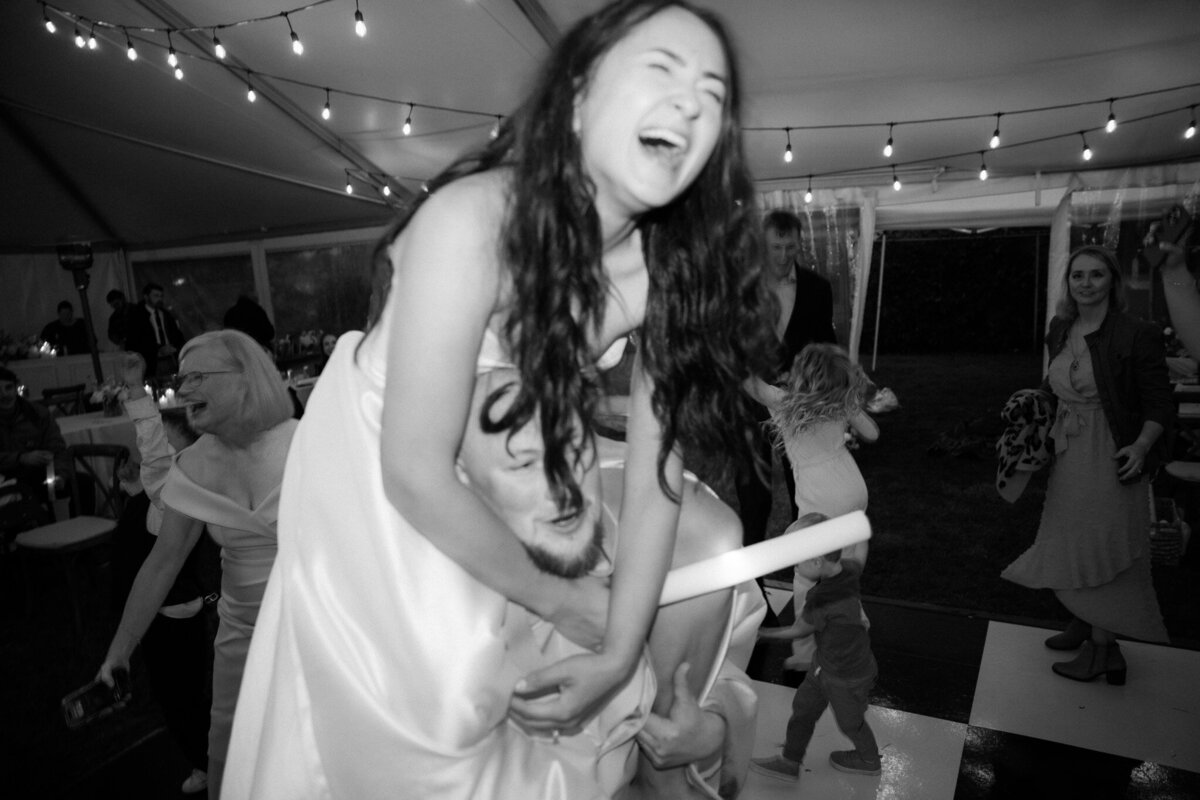 Bride dances on grooms shoulders at wedding under a lit tent in Oregon. 