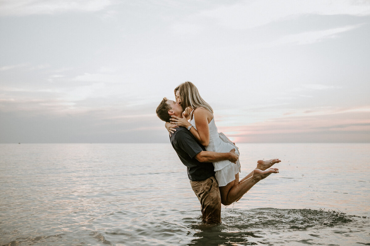 Fully clothed man in knee-deep water lifting up his fiance. Her feet are kicked up behind her and arms wrapped around his neck. They are kissing at golden hour.