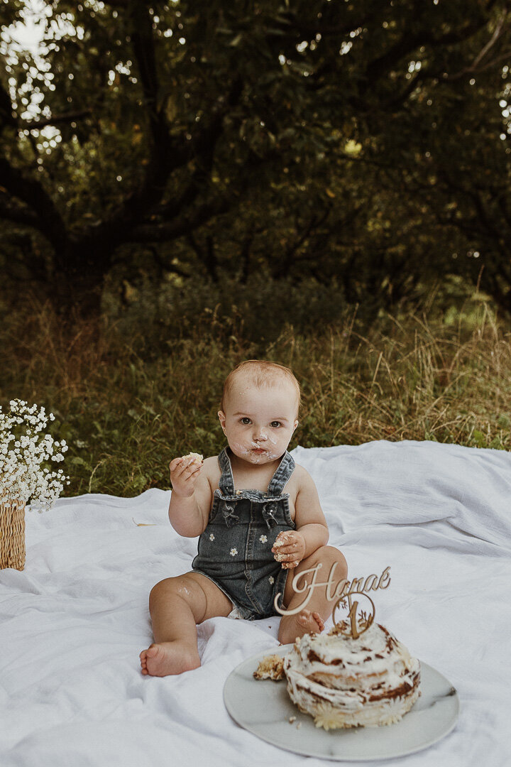 Photo d'une petite fille en salopette en jean à fleur pour ses un an. Séance smash the cake au milieu d'un champs après une averse d'été