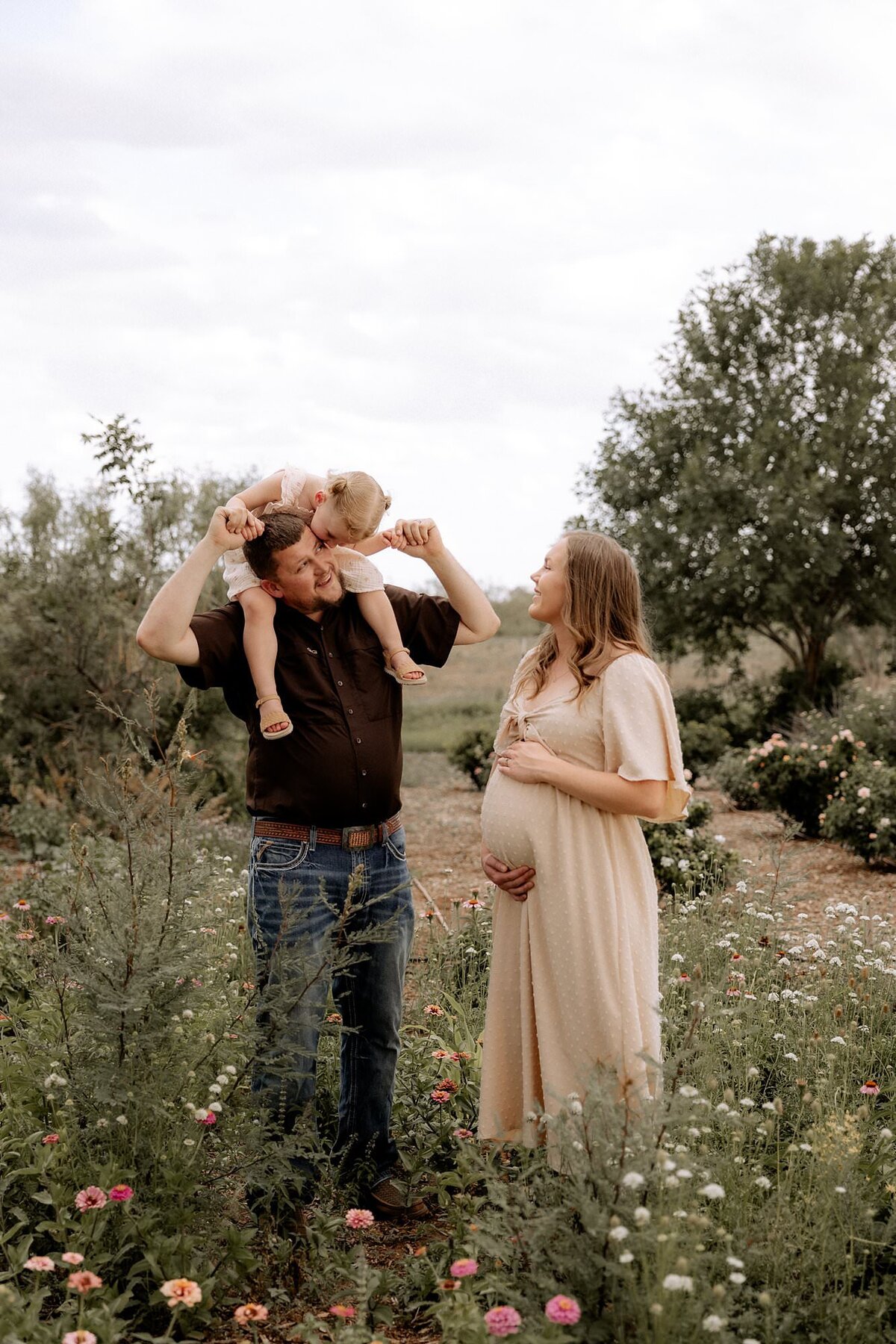dad has daughter sitting on his shoulder with pregnant wife at his side