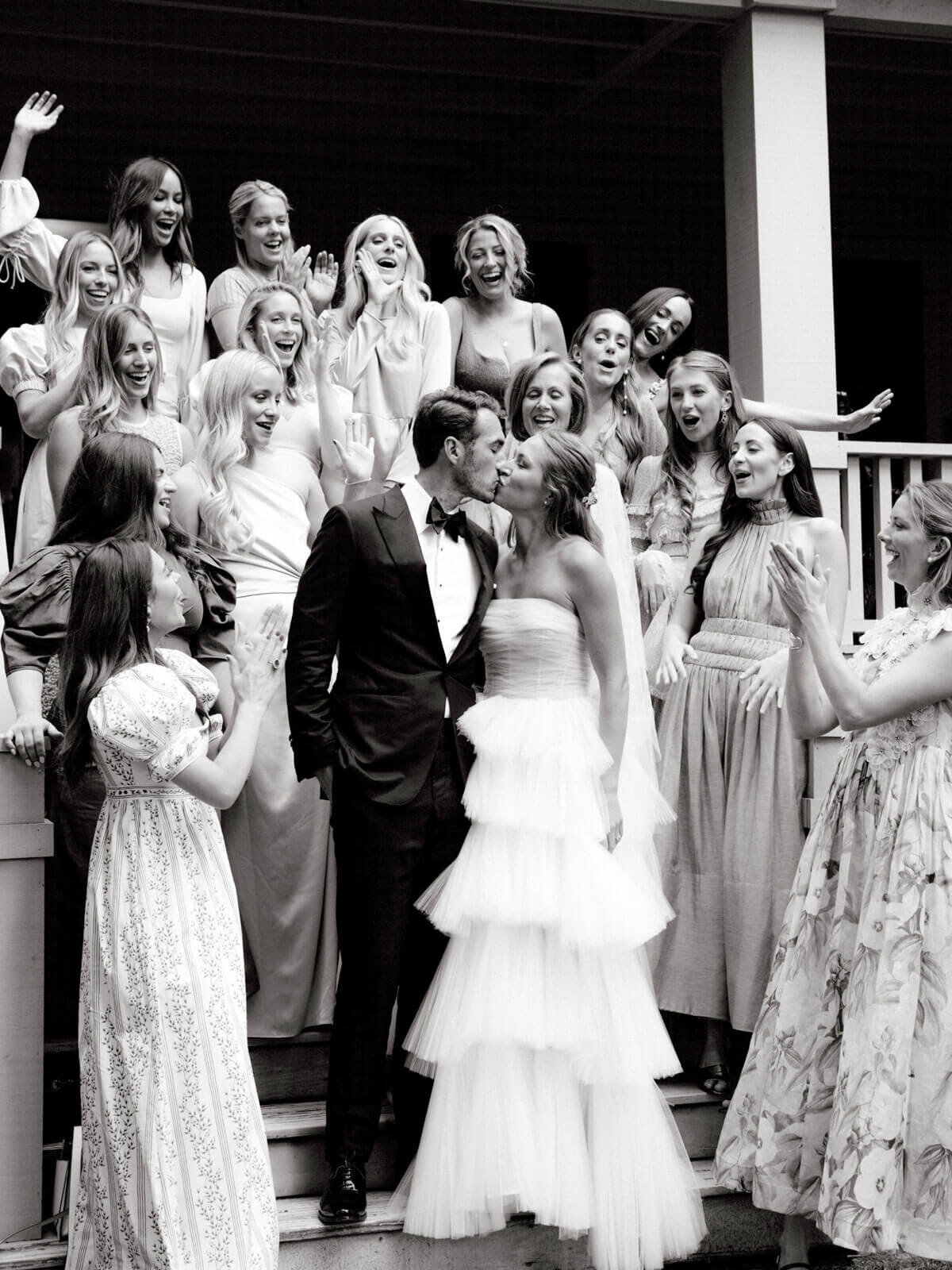 Black and white photo of the bride and groom kissing as the bridesmaids cheer on a staircase of a porch at The Ausable Club.