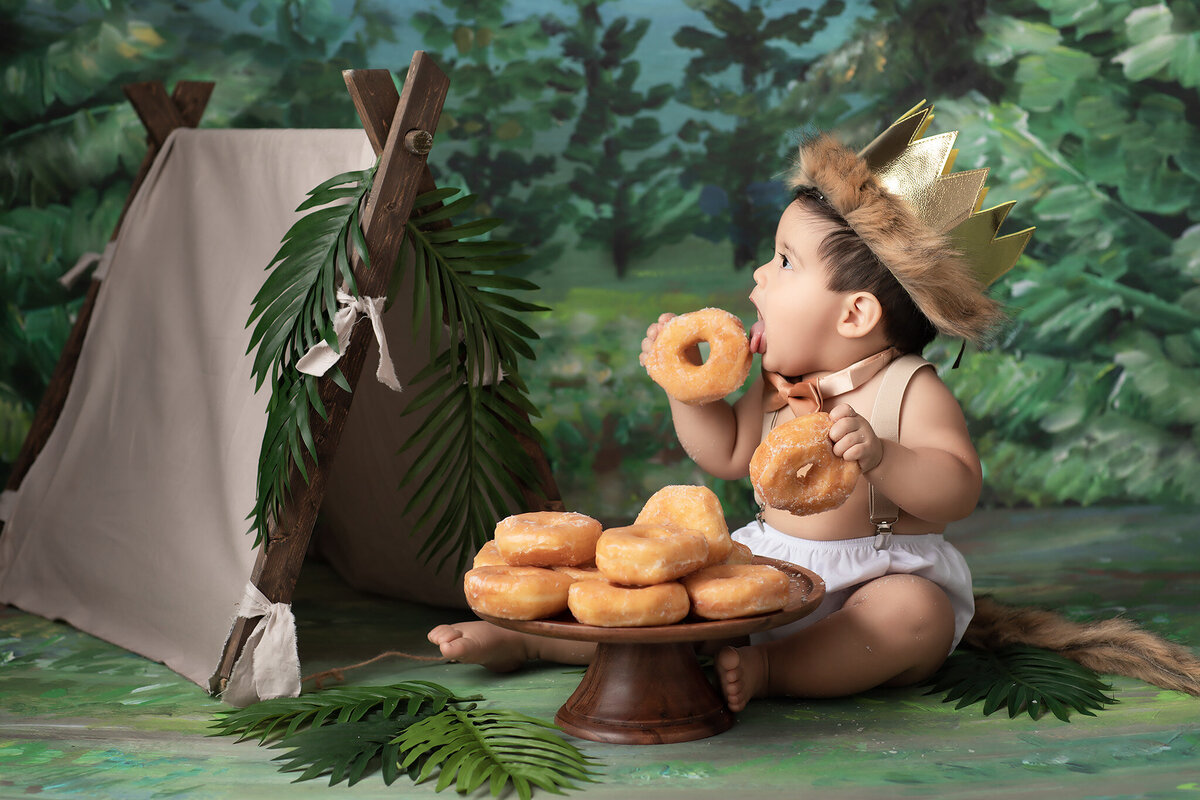 A playful baby wearing a gold crown with fur trim and a bow tie enjoys a pile of glazed donuts during a woodland-themed milestone photoshoot. Captured by a Dallas baby milestone photographer, this charming setup showcases the child's delight and curiosity in a fun and imaginative way.