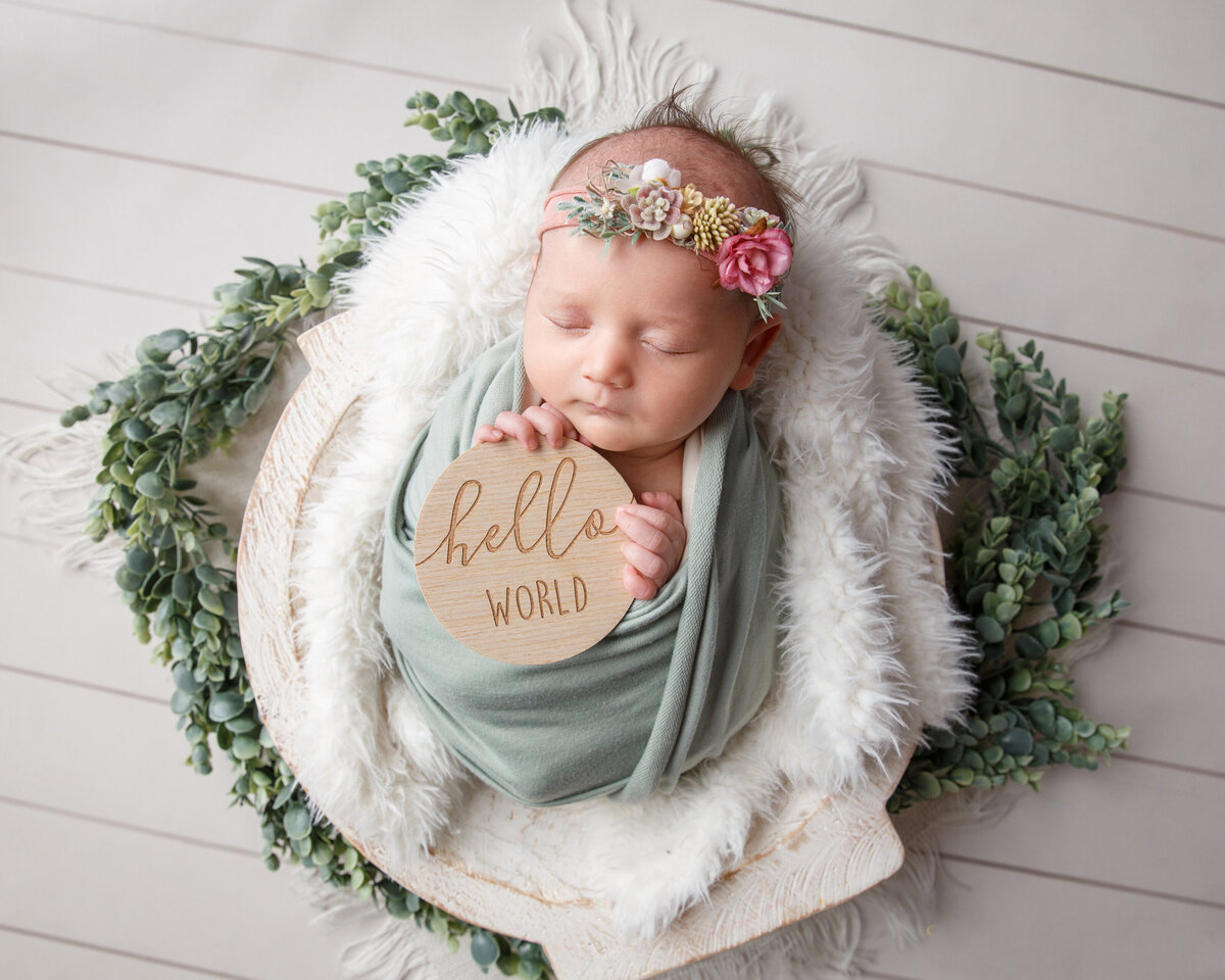 Newborn baby girl wearing a flowered grown and wrapped in a light green scarf holding a hello world sign