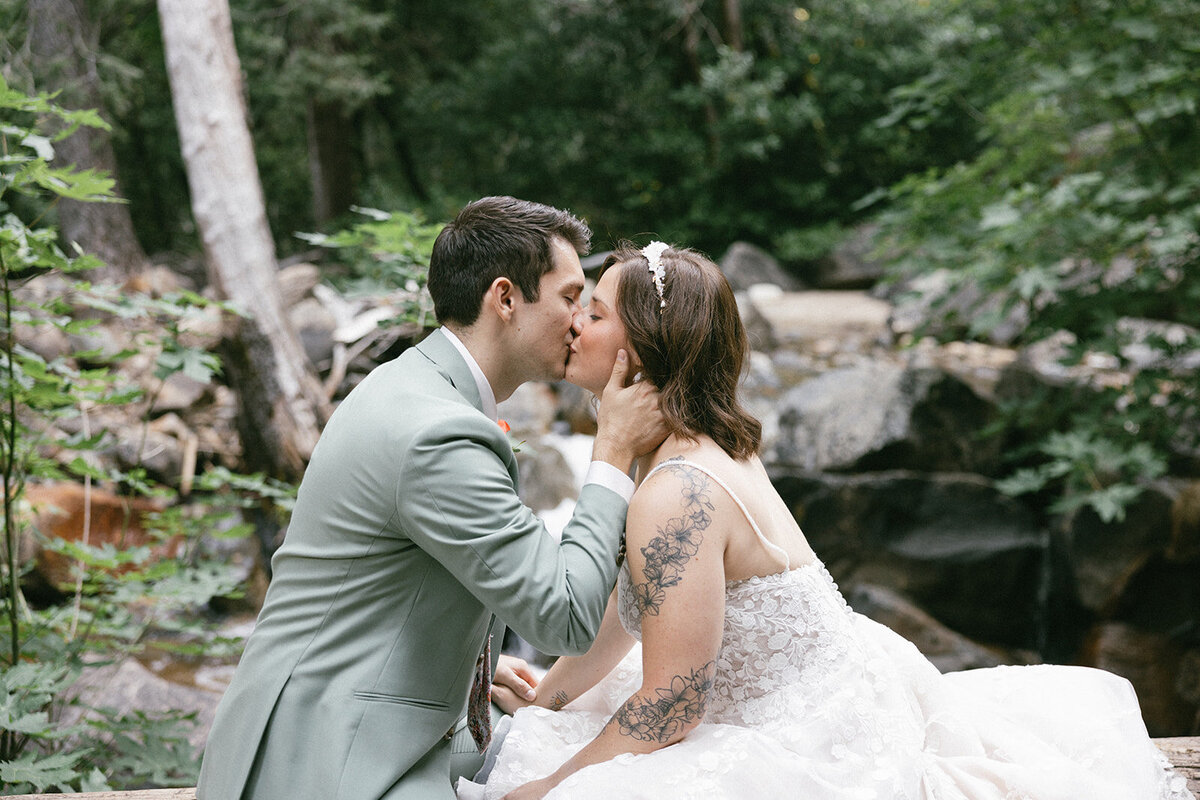 Bride and groom in wedding attire casually sit on a log in the forest of Yosemite while sharing a kiss. 