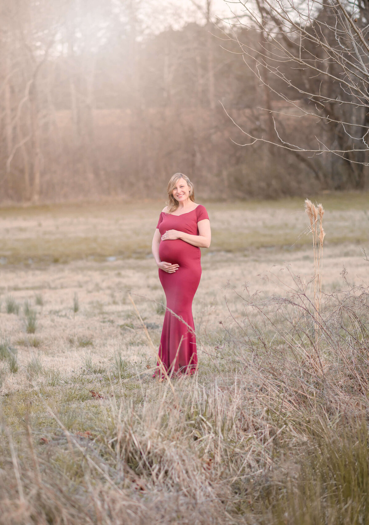 beautiful mom to be on a field during fall  season, smiling at the camera while hugging her 32 weeks pregnant belly