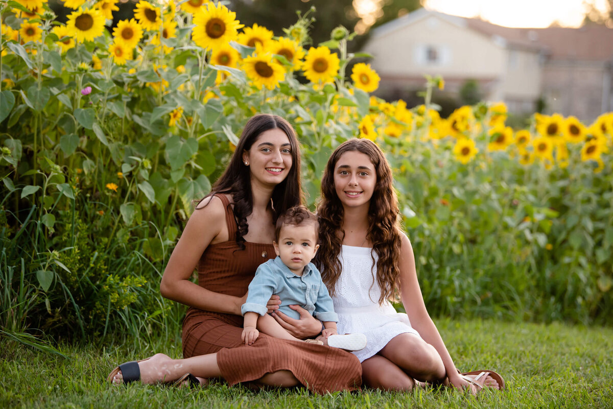 Teen sisters sitting on the ground with their baby brother in front of a sunflower field for their family photos
