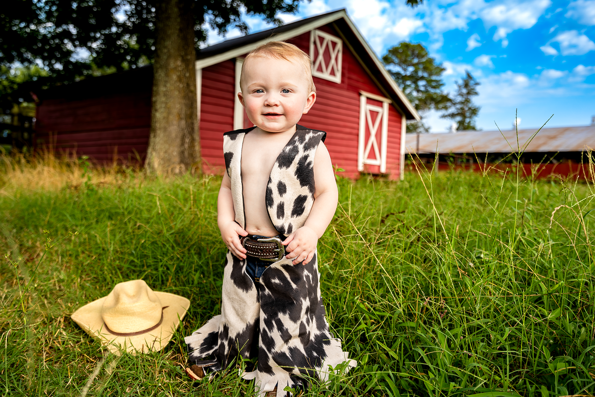 1 year old cowboy in front of a red barn under blue skies