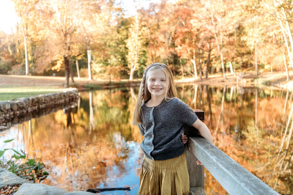 Connecticut family photographer documents young girl in mustard skirt posing by pond with beautiful autumn foliage reflections