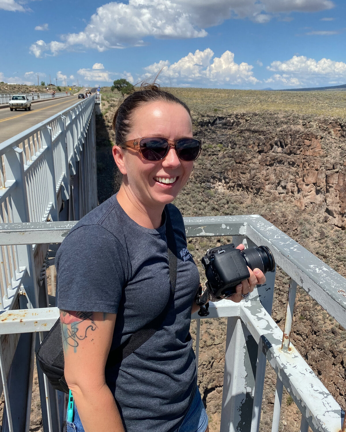 woman standing on a bridge overlooking a rock valley, holding her camera, wearing sunglasses and smiling at the camera