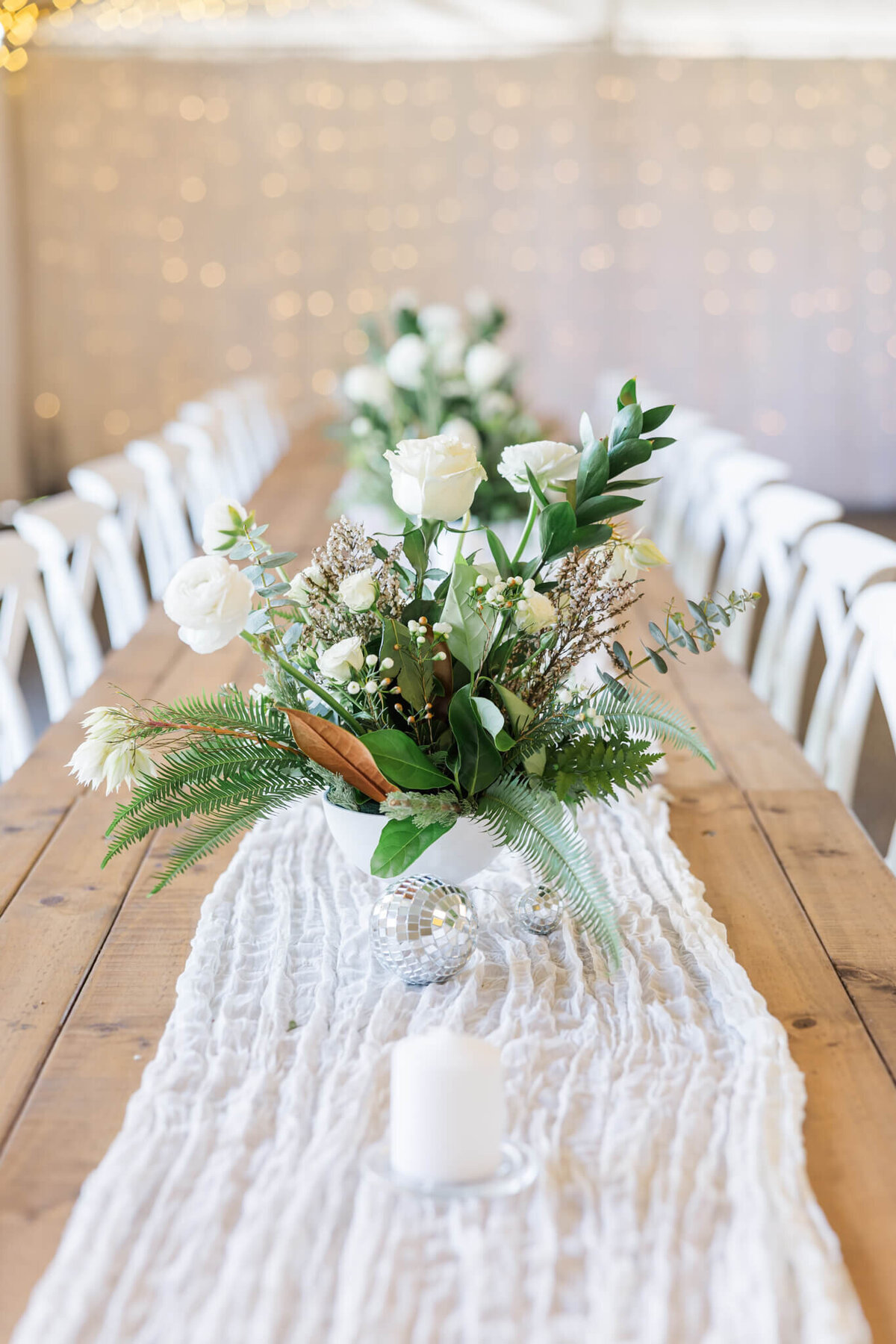 White and cream florals surrounded by green ferns and leaves in a flower arrangement on a wedding reception table. 