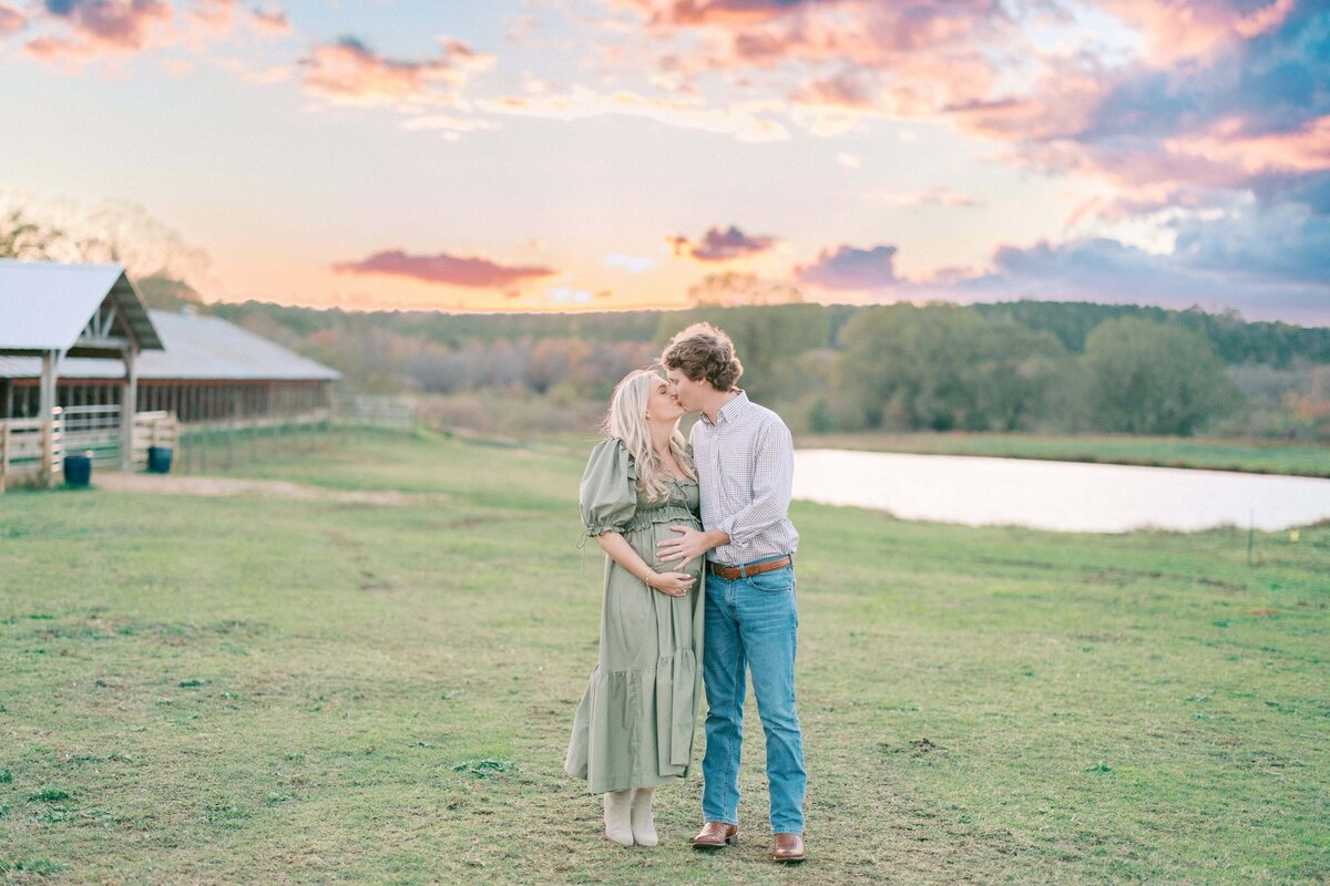 Pregnant woman in green dress kisses husband in green field with sunset skies