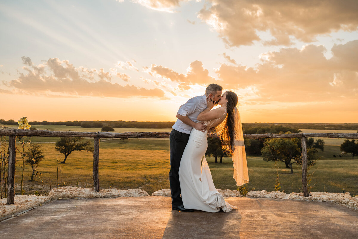 Groom dips bride and kisses her at sunset overlooking ranch.