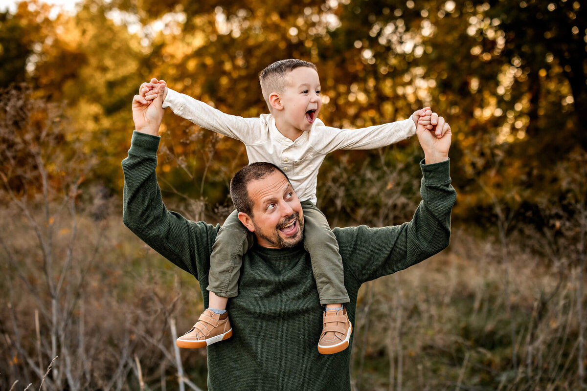 A man with a beard, wearing a green sweater, carries a smiling young boy on his shoulders in an outdoor portrait. The boy, dressed in a light long-sleeve shirt and green pants, joyfully holds the man's hands, set against the backdrop of autumn trees captured by a Bloomington family photographer.