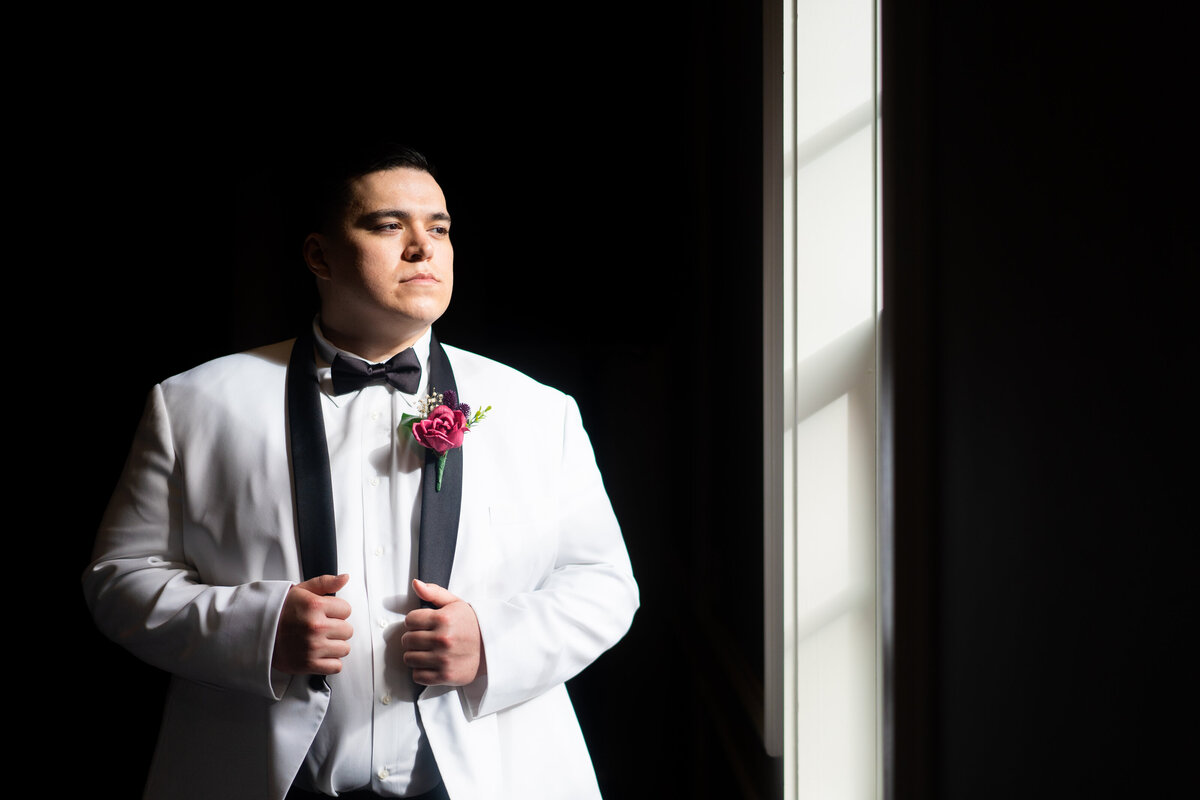 Groom in a white tux standing in direct sunlight looks out the window at his wedding at Nationwide Hotel and Conference Center in Lewis Center, Ohio.