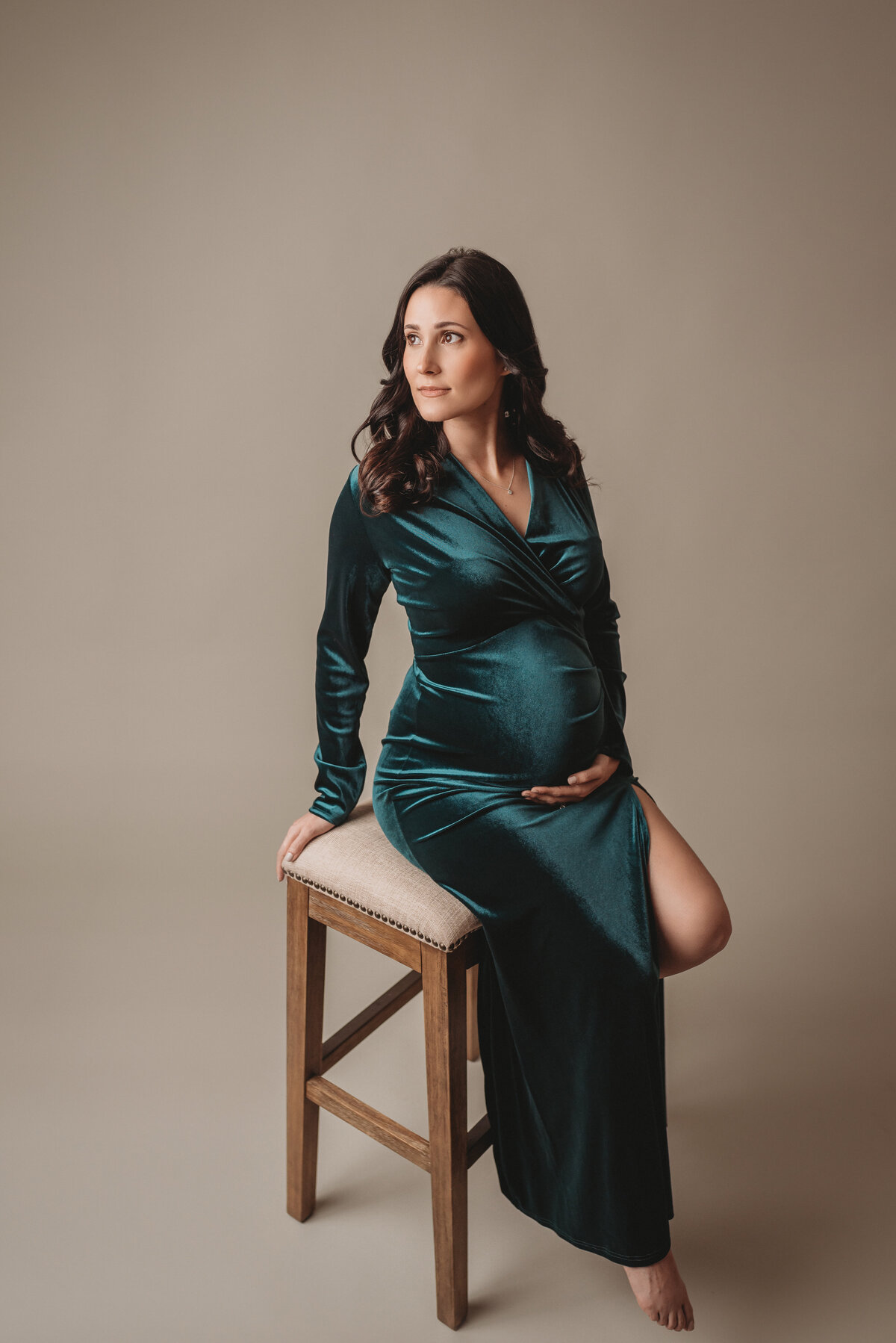 Pregnancy portrait of woman wearing green velvet gown sitting on a stool holding baby bump