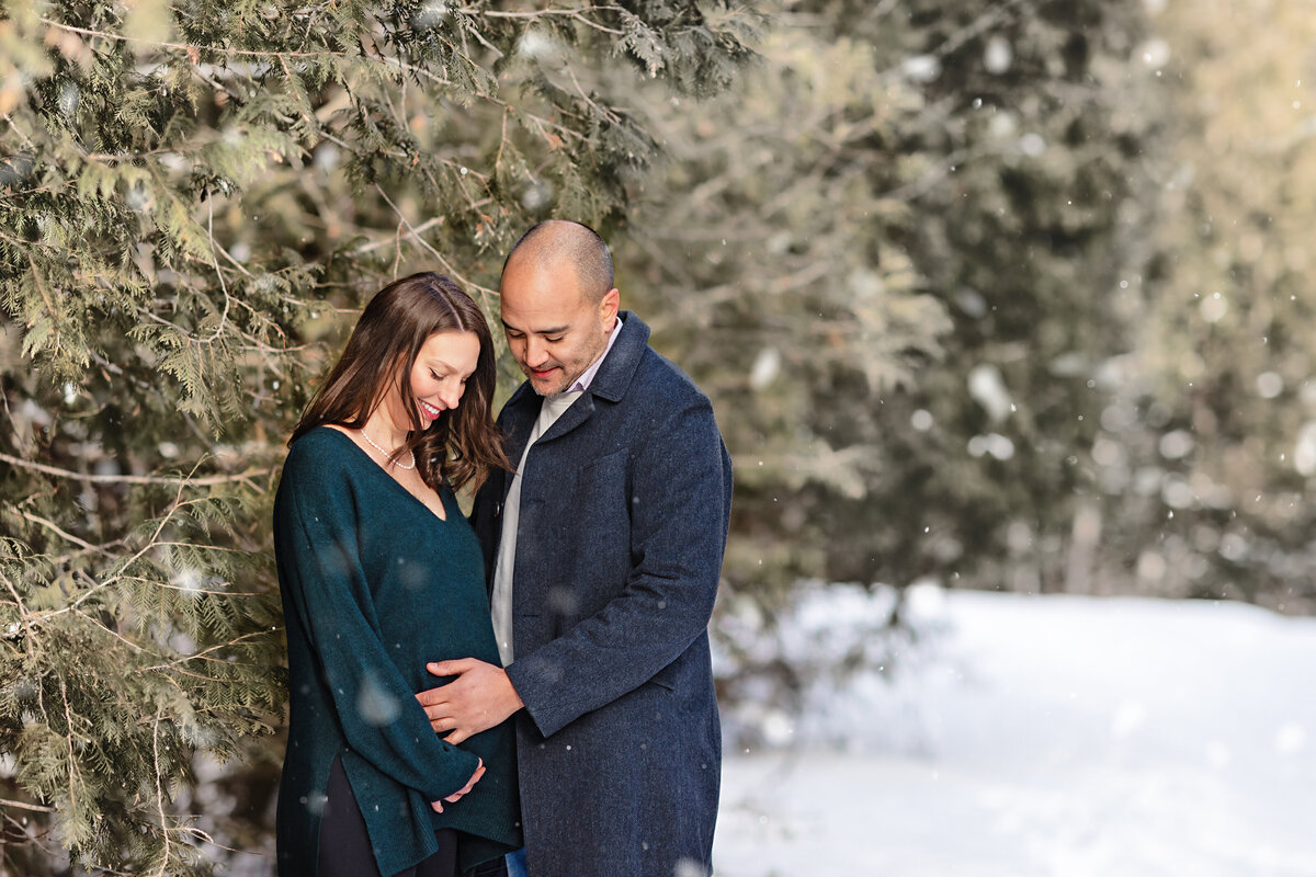 Expecting couple sharing a tender moment in a snowy pine forest during their winter maternity session in Paris, Ontario