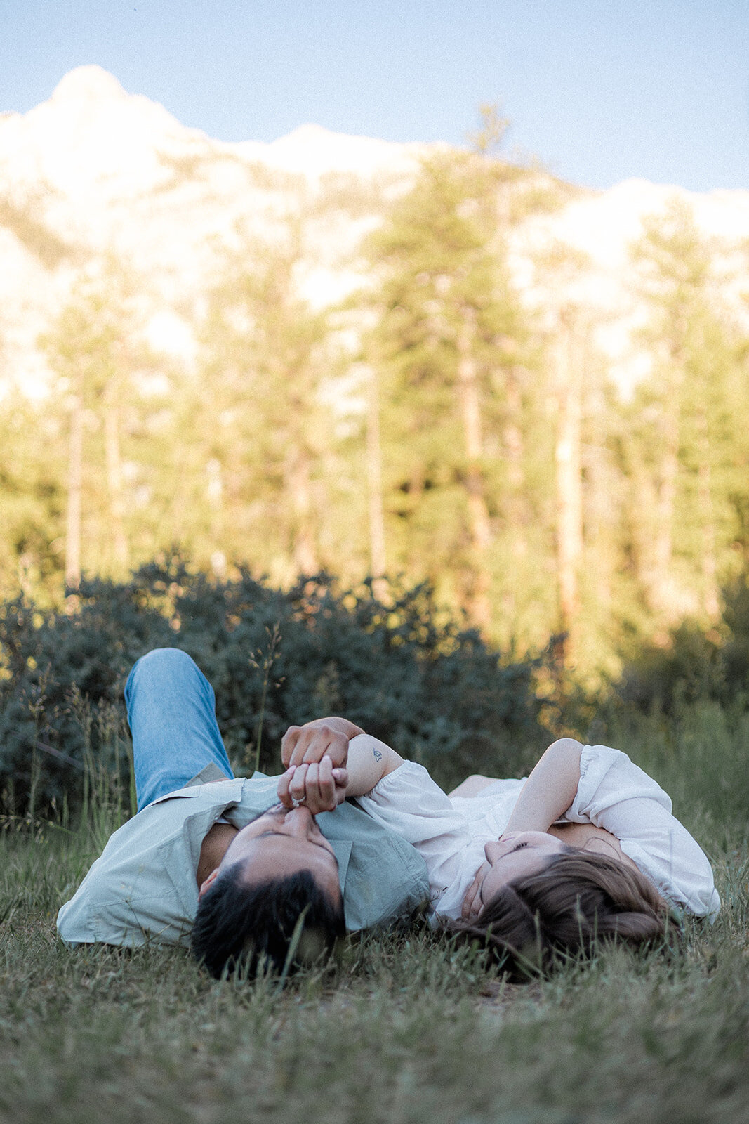The couple lies side by side in the grass, holding hands and looking relaxed, surrounded by greenery and distant mountain views.