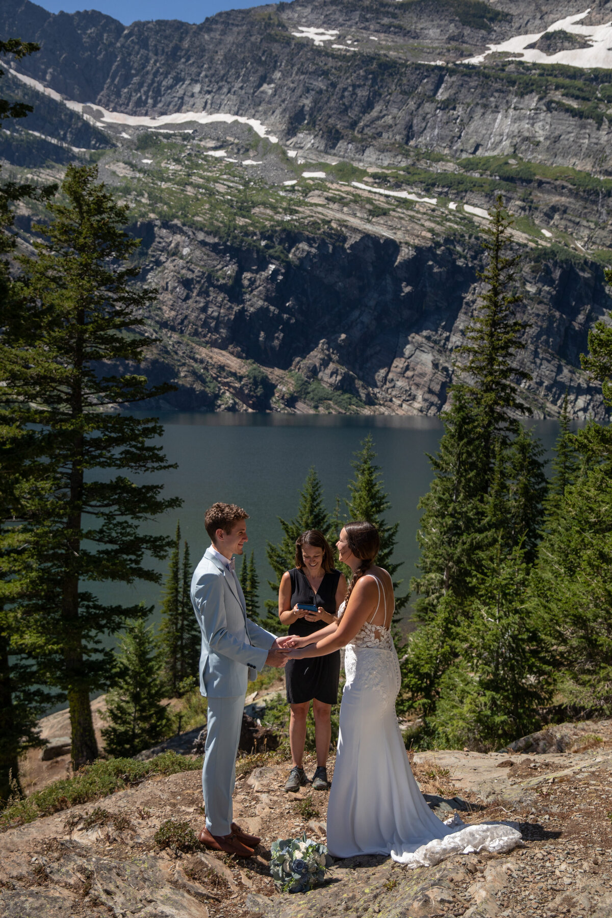 A couple stand facing each other and holding hands with their officiant standing behind them and snow capped mountains and a lake behind her.