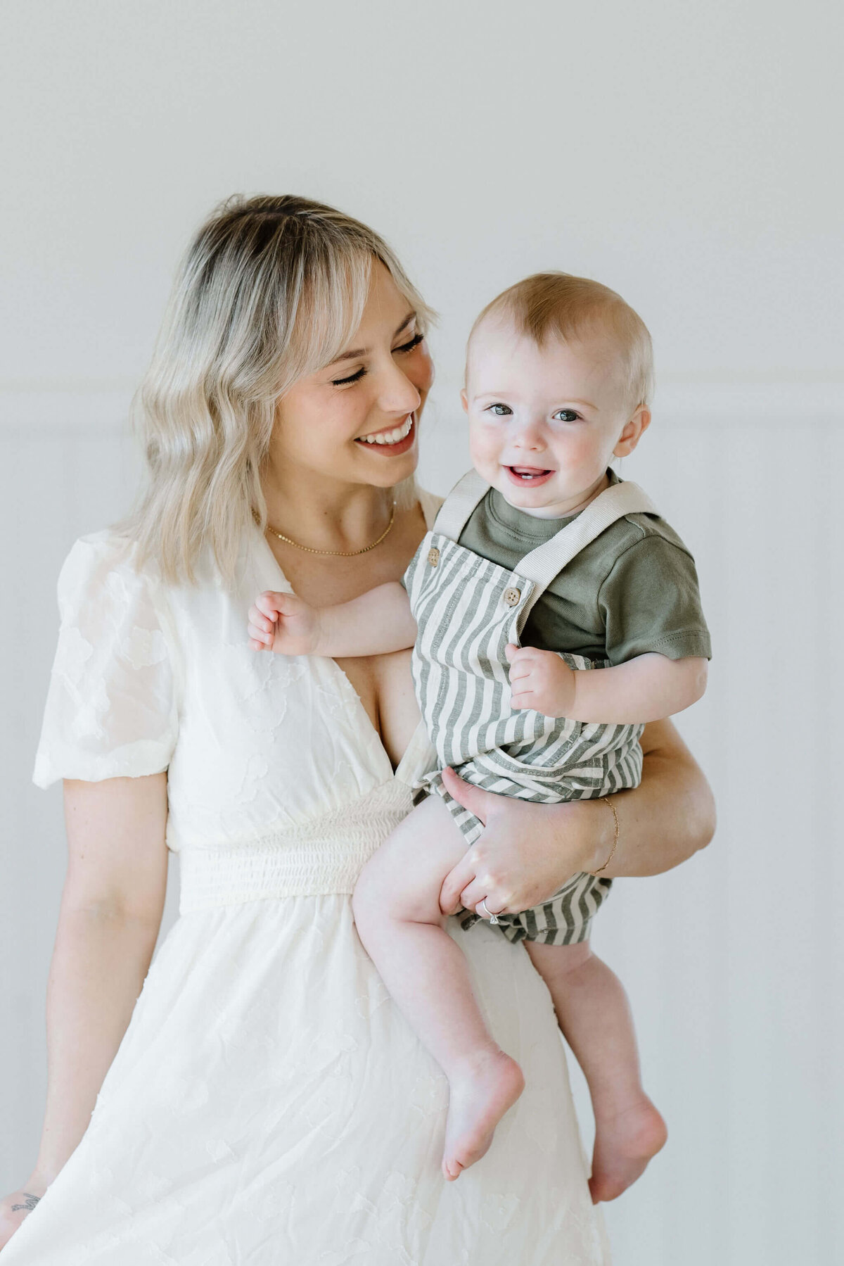 mother in white dress holding one year old son in stripped overalls and green shirt