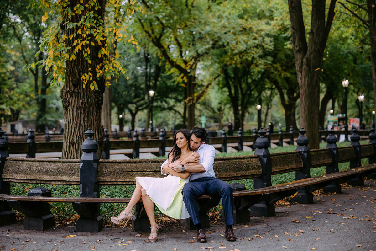 The fiancé is wrapping his arms around his fiancée as they sit on the bench at Central Park, NYC. Image by Jenny Fu Studio.