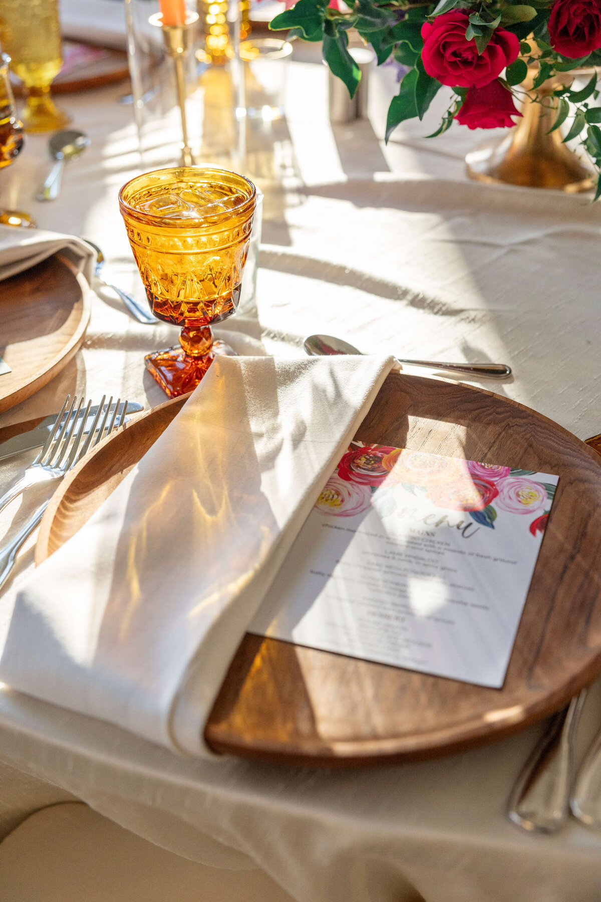 A table setting with a wooden plate holding a white napkin and a floral menu card. An amber glass goblet is next to the plate. Sunlight casts shadows over the white tablecloth adorned with flowers and silverware.